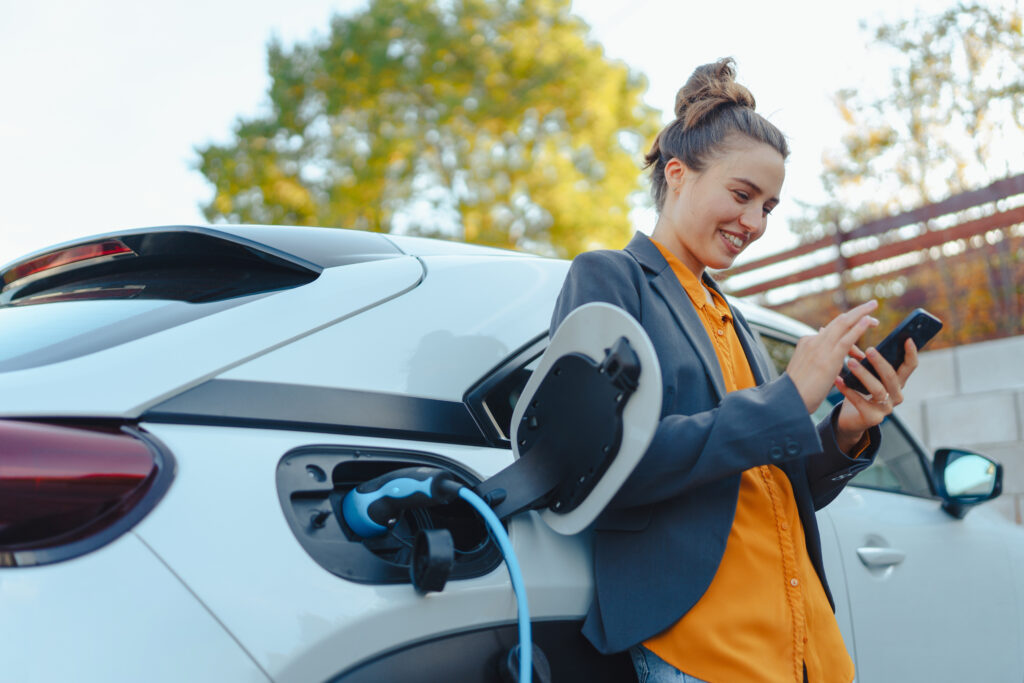 An female EV driver views details of an active charging session on their mobile phone while leaning against their electric car, which is plugged in and charging